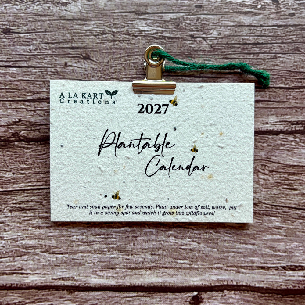 A 2027 plantable calendar card by A La Kart Creations, held by a gold metal clip with a green string against a rustic wood plank background.