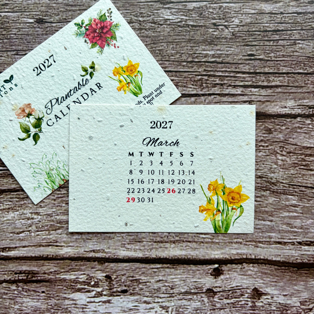 Two plantable paper calendar cards with floral illustrations are arranged on a rustic weathered wood surface.
