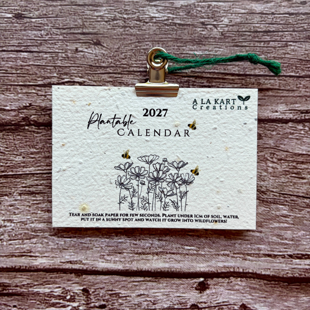 A 2027 plantable paper calendar card by La Kart Creations, clipped with a gold metal clip and green string, displayed on a rustic brown wood background.