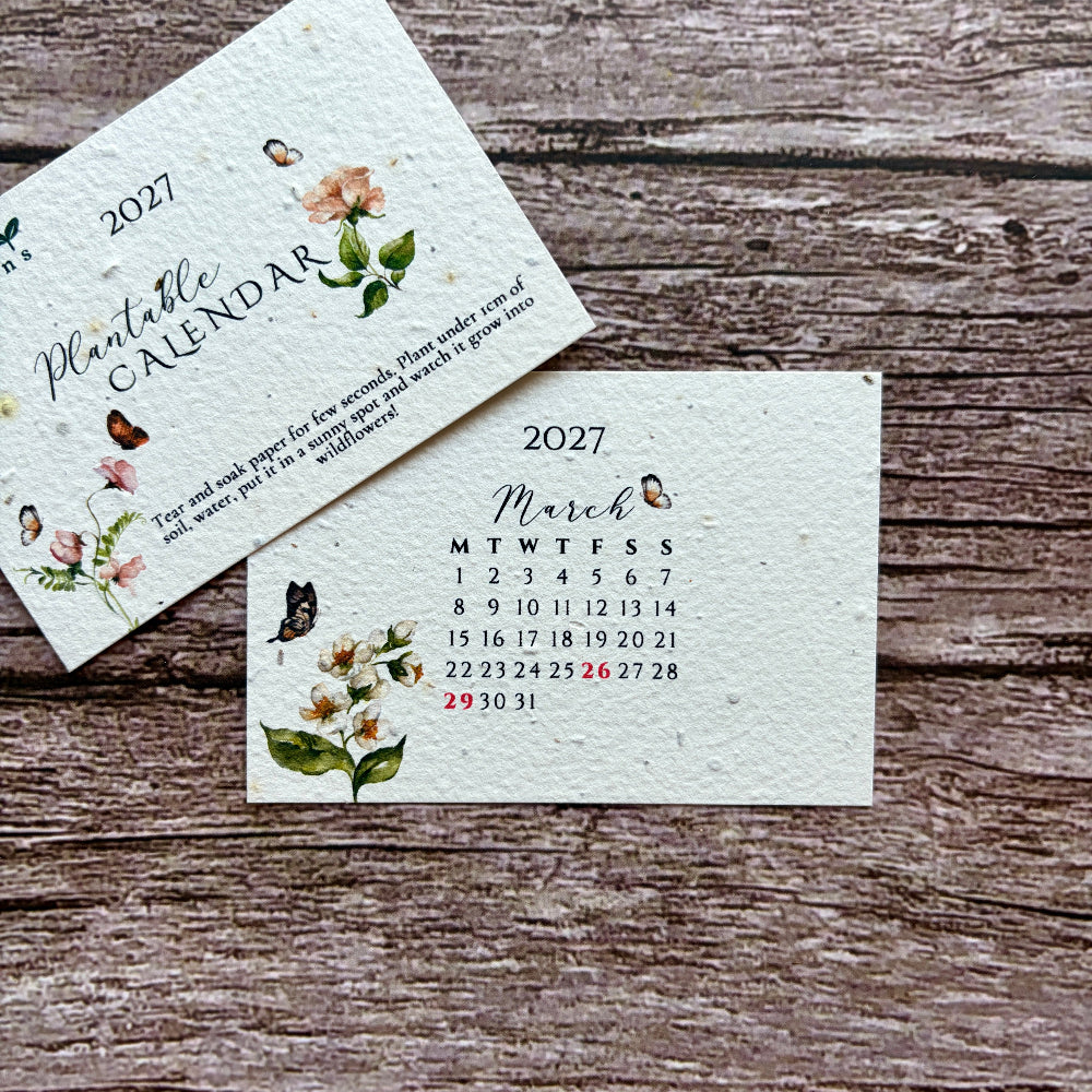 Two small 2027 plantable calendar cards with floral illustrations and butterflies, displayed on a rustic wooden surface. One card shows the full calendar cover and the other shows the March monthly layout.