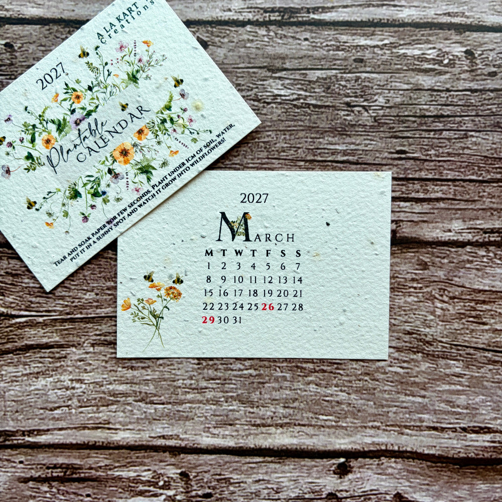 A 2027 floral monthly calendar card and its cover shown on a rustic wooden surface. The cover features delicate watercolor-style yellow, orange, and green flowers with the title &