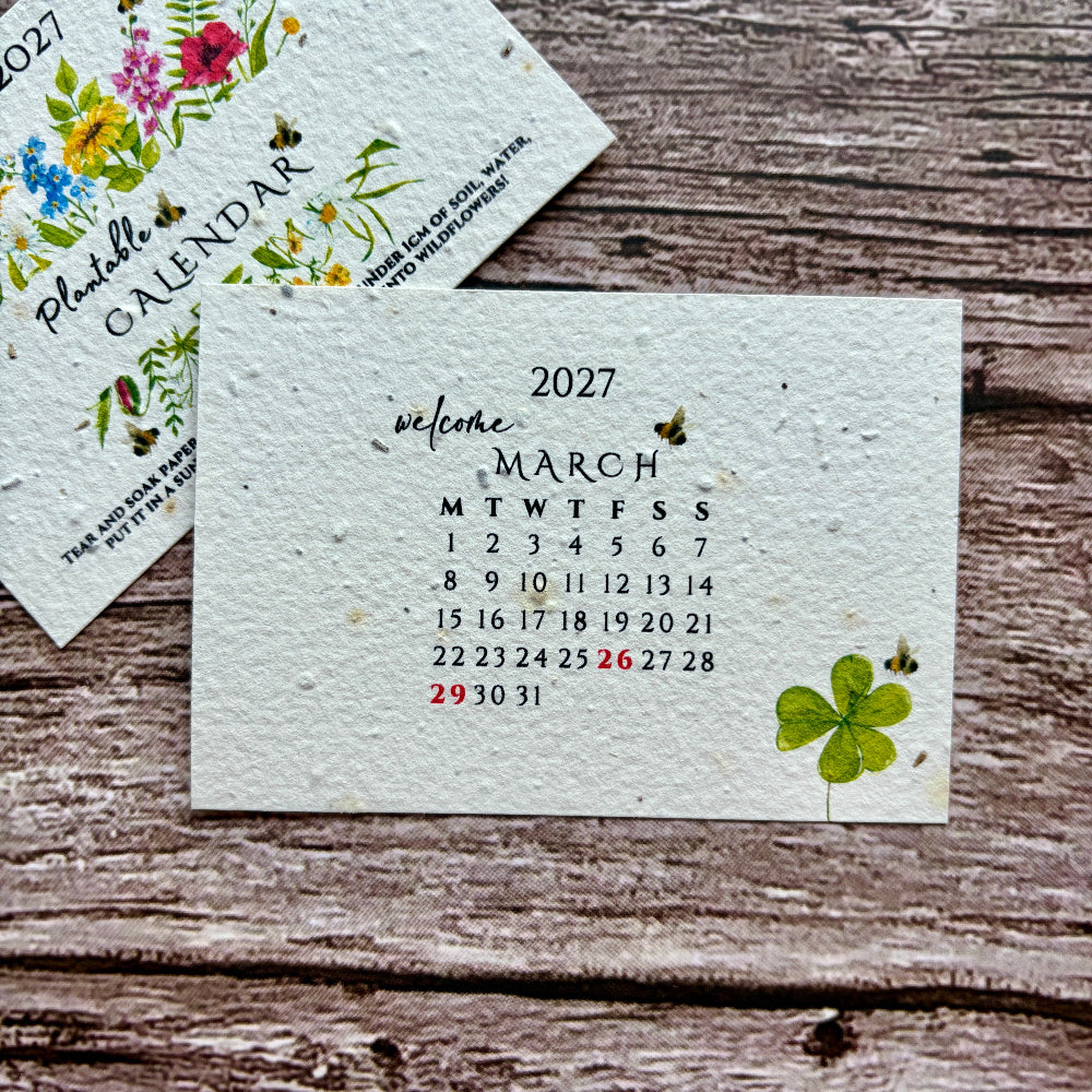 A handmade seed paper 2027 monthly calendar card with watercolor floral and butterfly illustrations, displayed on a rustic wooden background next to another calendar page.