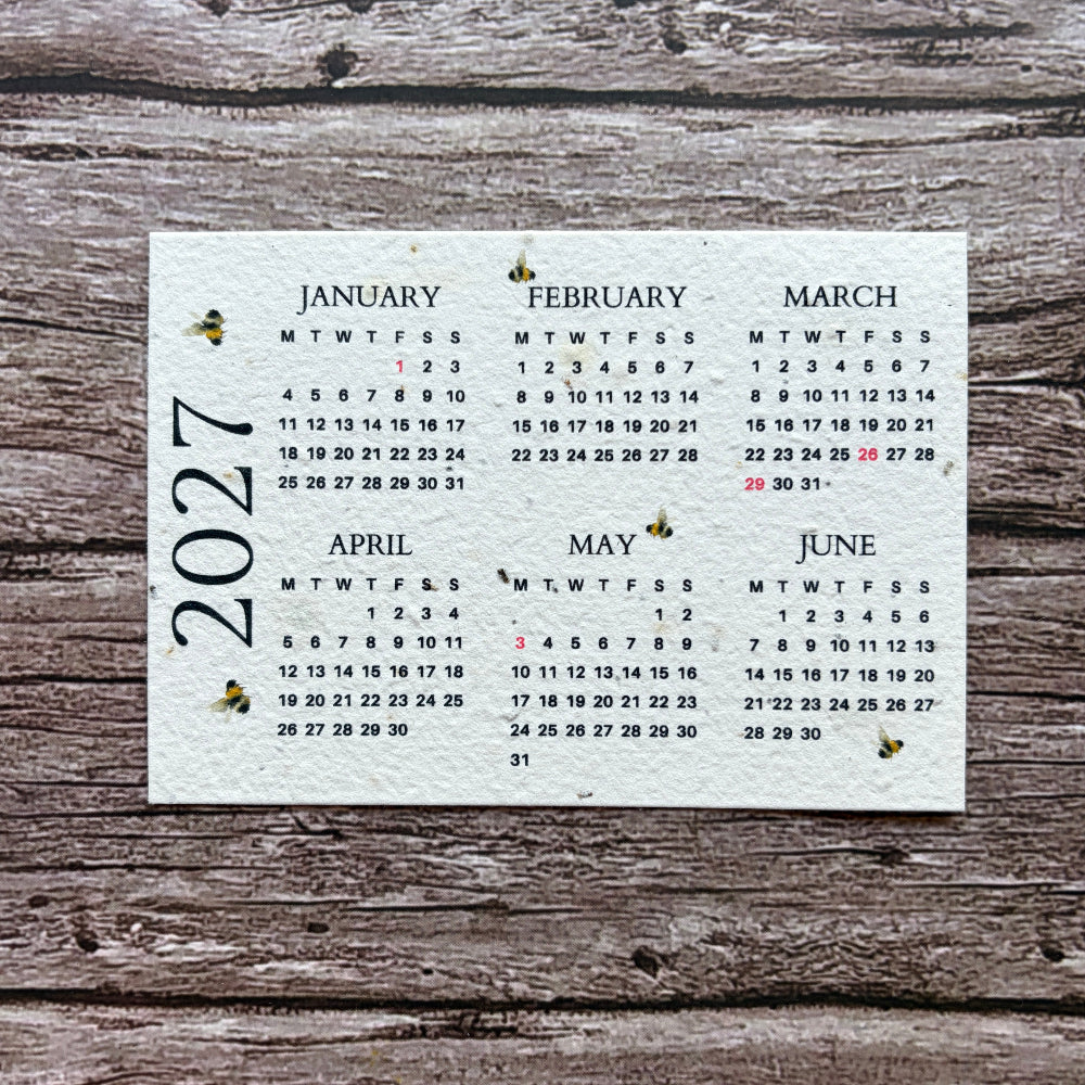 A square white seed paper calendar page showing the months of 2027 in black text, placed on a rustic weathered wood surface.