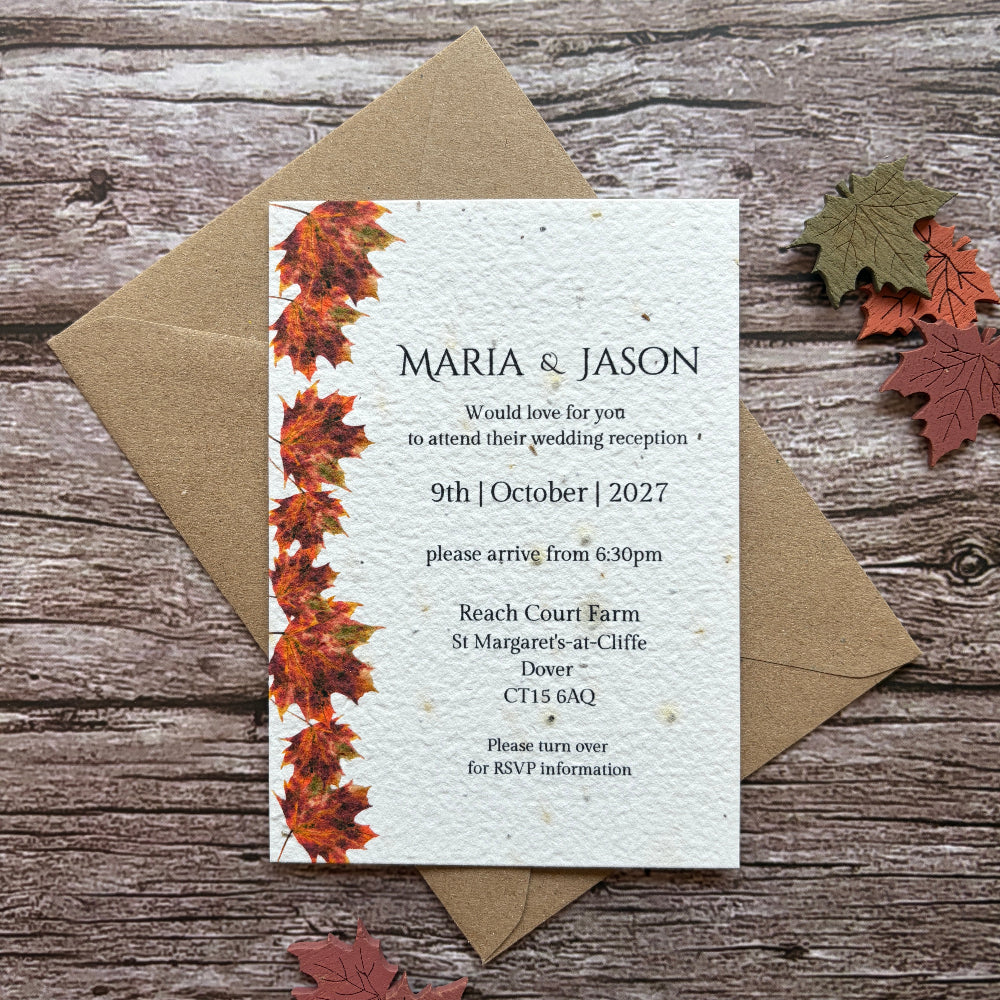 A plantable wedding invitation card with a vertical border of orange and red autumn leaves, placed on top of a brown kraft envelope on a rustic wood background with scattered fall leaves.