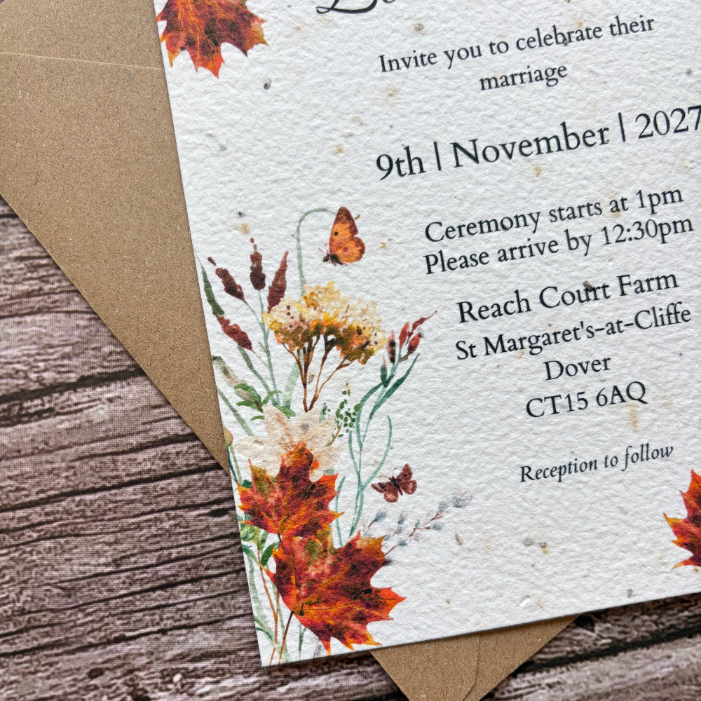 A rustic fall-themed wedding invitation card with watercolor autumn leaves and wildflowers, placed on top of a kraft envelope over a weathered wood surface.