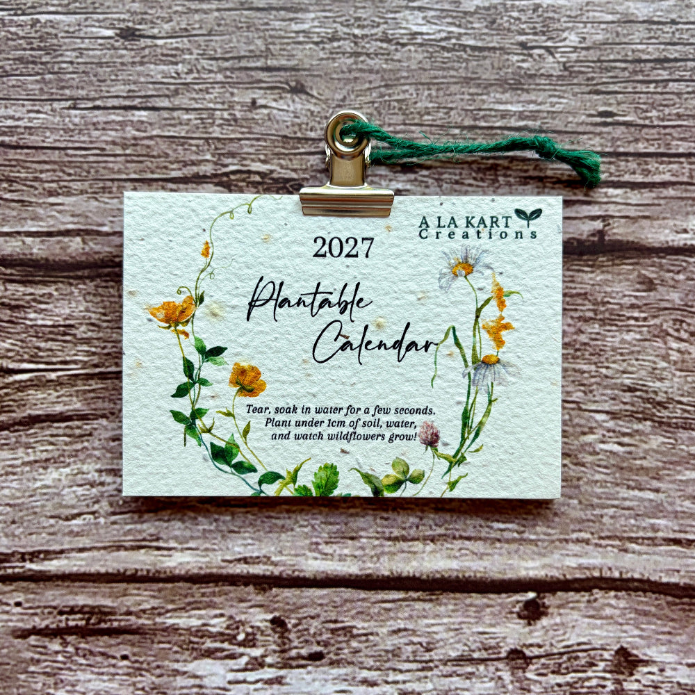 A 2027 Plantable Calendar card by A La kart Creations, featuring a watercolor floral wreath design, held by a gold clip with a green string against a rustic wooden background.
