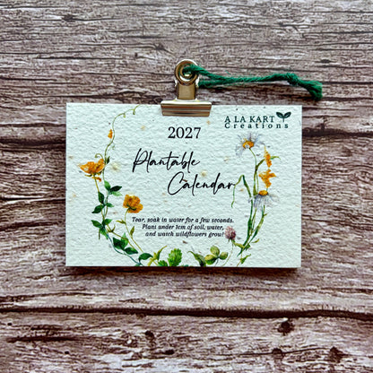 A 2027 Plantable Calendar card by A La kart Creations, featuring a watercolor floral wreath design, held by a gold clip with a green string against a rustic wooden background.