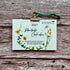 A 2027 Plantable Calendar card by A La kart Creations, featuring a watercolor floral wreath design, held by a gold clip with a green string against a rustic wooden background.