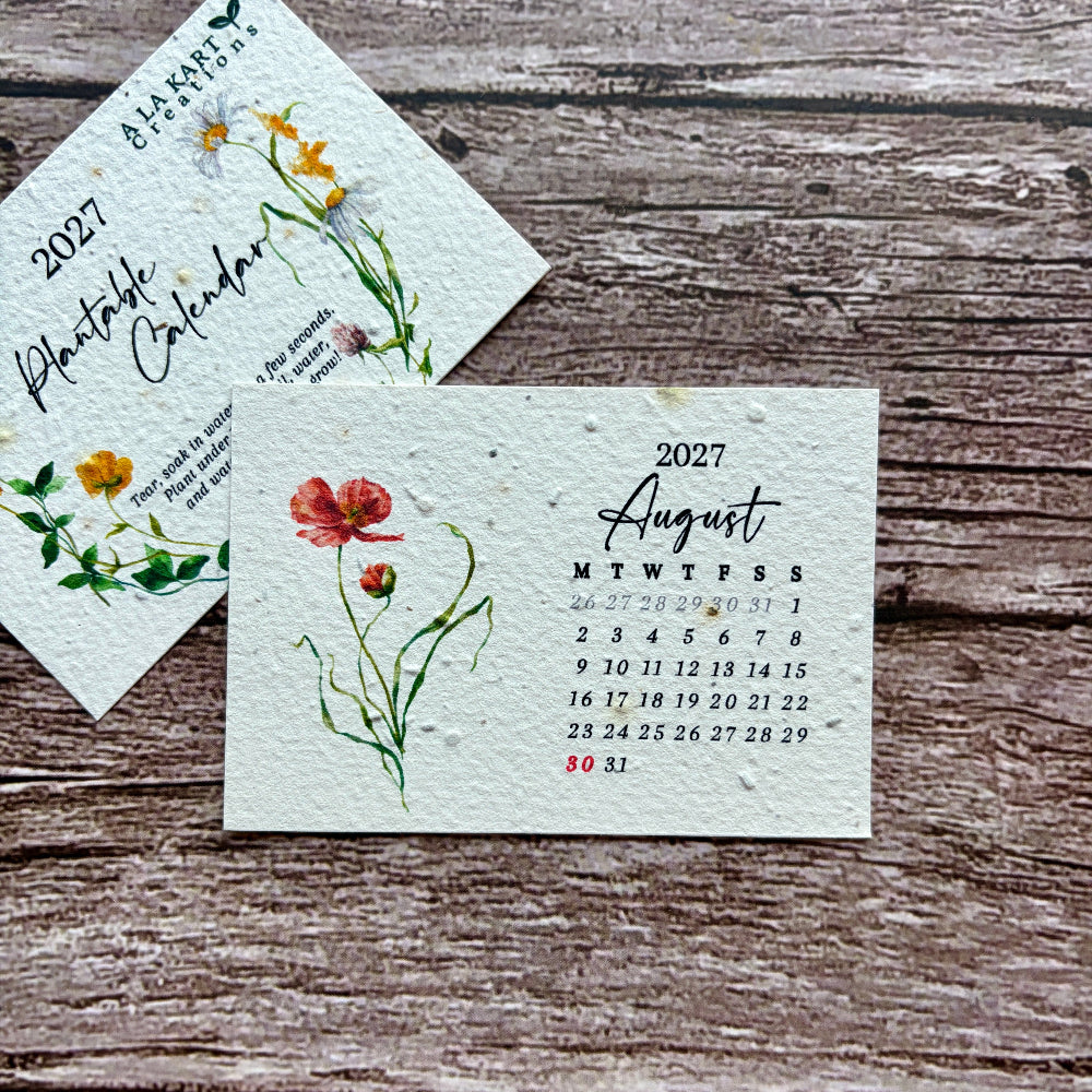 Two square paper plantable calendar cards with watercolor floral illustrations are arranged on a rustic wooden surface. One card shows the full 2027 calendar, and another partially visible card shows the August 2027 page.