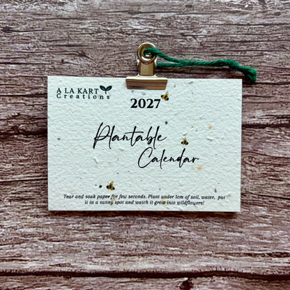 A 2027 plantable calendar card by A La Kart Creations, held by a gold metal clip with a green string against a rustic wood plank background.