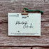 A 2027 plantable calendar card by A La Kart Creations, held by a gold metal clip with a green string against a rustic wood plank background.