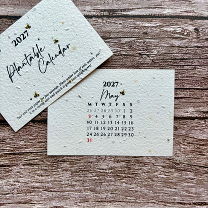 Two small square paper cards on a rustic wood surface. One card reads "2027 Plantable Calendar" and the other shows the May 2027 calendar dates.