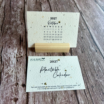 A small 2027 monthly plantable calendar card set displayed on a rustic wood background, with one card standing upright on a wooden block and the other lying flat showing the cover design.