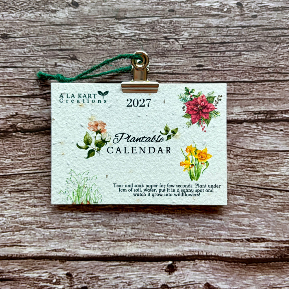 A 2027 plantable paper calendar card by A La KArt Creations featuring watercolor floral illustrations and a green hanging ribbon, displayed on a rustic weathered wood background.