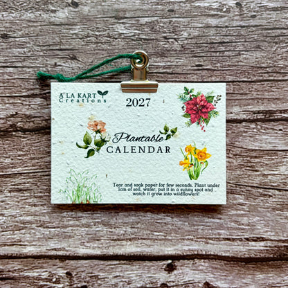 A 2027 plantable paper calendar card by A La KArt Creations featuring watercolor floral illustrations and a green hanging ribbon, displayed on a rustic weathered wood background.