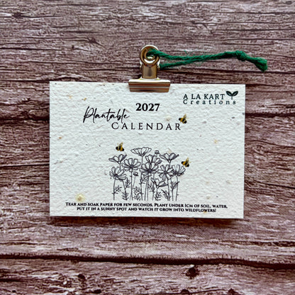 A 2027 plantable paper calendar card by La Kart Creations, clipped with a gold metal clip and green string, displayed on a rustic brown wood background.
