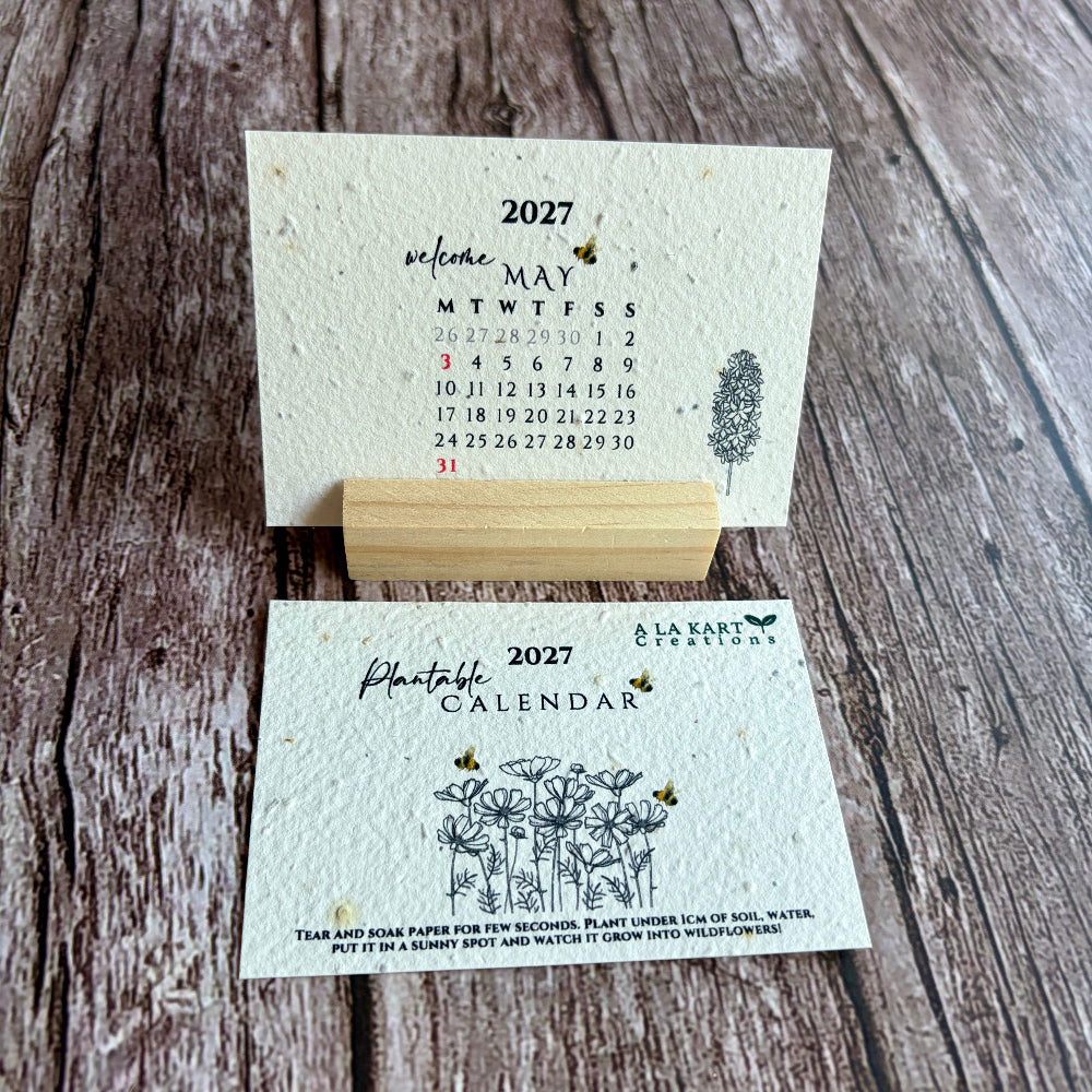 A handmade 2027 paper calendar displayed on a rustic wood background, featuring a front cover with wildflower illustrations and a back cover showing the monthly calendar layout.