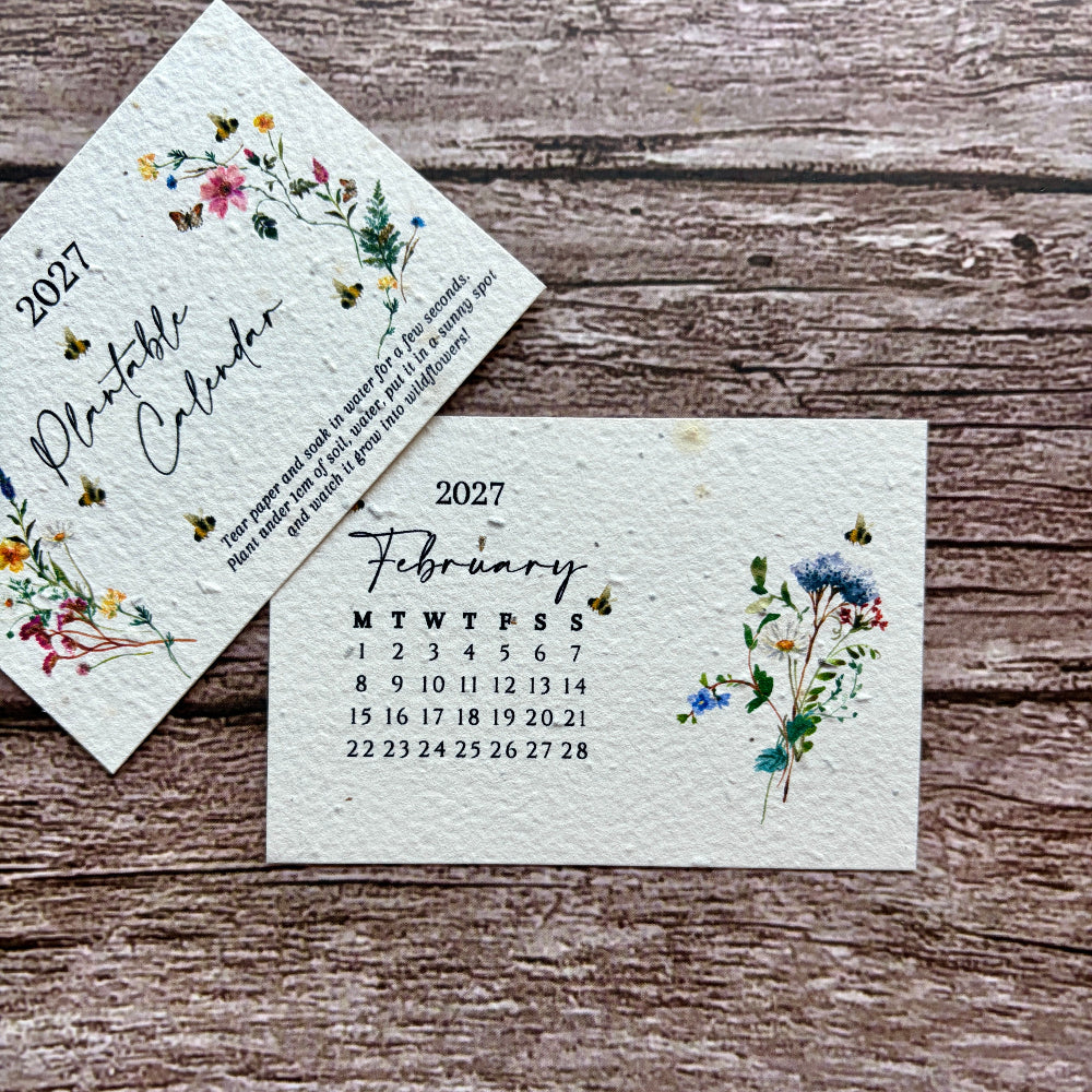 A handmade seed paper monthly calendar page for February 2027 with floral watercolor illustrations, displayed on a rustic wooden surface next to another angled page showing the year 2027.