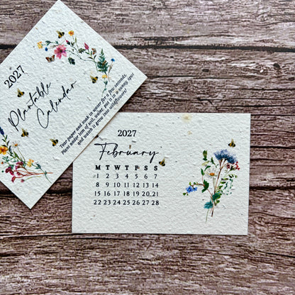 A handmade seed paper monthly calendar page for February 2027 with floral watercolor illustrations, displayed on a rustic wooden surface next to another angled page showing the year 2027.