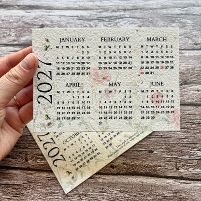 A hand holds a printed 2027 plantable monthly calendar card over another overlapping calendar card, set against a rustic wood plank background.