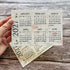 A hand holds a printed 2027 plantable monthly calendar card over another overlapping calendar card, set against a rustic wood plank background.