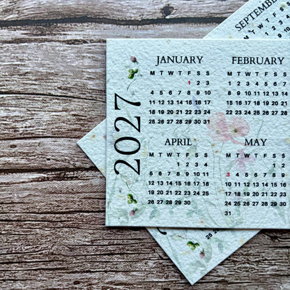 A set of printed monthly plantable calendar cards for 2027, including January through September, arranged on a rustic wooden surface. The cards are light blue with black text and small pink floral accents.