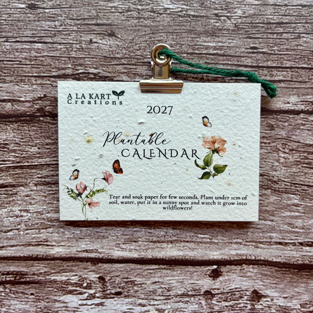 A 2027 Plantable Calendar by A La Kart Creations, made from textured watercolor paper with floral illustrations and a green hanging string, clipped to a rustic wood background.