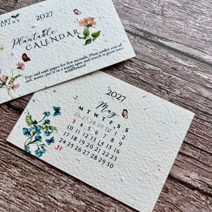 Two handmade paper plantable calendar cards for 2027, one showing the cover with floral illustrations and the other showing the monthly calendar layout for May, placed on a rustic wooden surface.