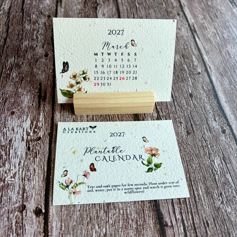 A 2027 plantable monthly calendar from A La Kart Creations displayed on a rustic wooden background with a small wooden block stand.