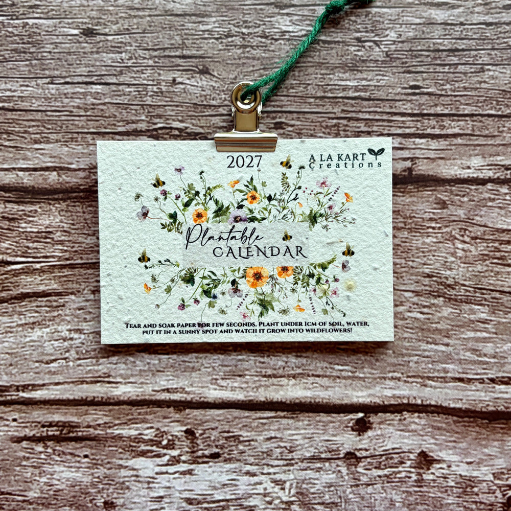 A 2027 monthly calendar by A La KArt Creations featuring a delicate watercolor floral design, held by a brass clip on a green cord, displayed flat on a rustic weathered wood background.