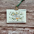 A 2027 monthly calendar by A La KArt Creations featuring a delicate watercolor floral design, held by a brass clip on a green cord, displayed flat on a rustic weathered wood background.