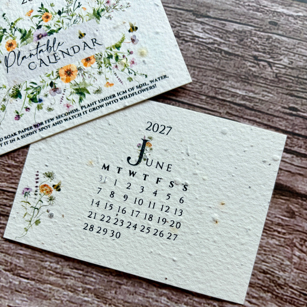 A close-up of a handmade plantable paper calendar page from 2027 showing June, placed on a rustic wooden surface next to another floral calendar card.