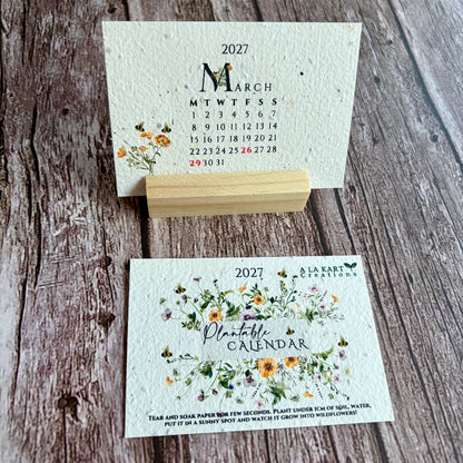 A handmade paper monthly calendar for March 2027 displayed on a small wooden stand, with the front cover showing floral illustrations and the back cover showing the monthly calendar layout, set on a rustic wood background.