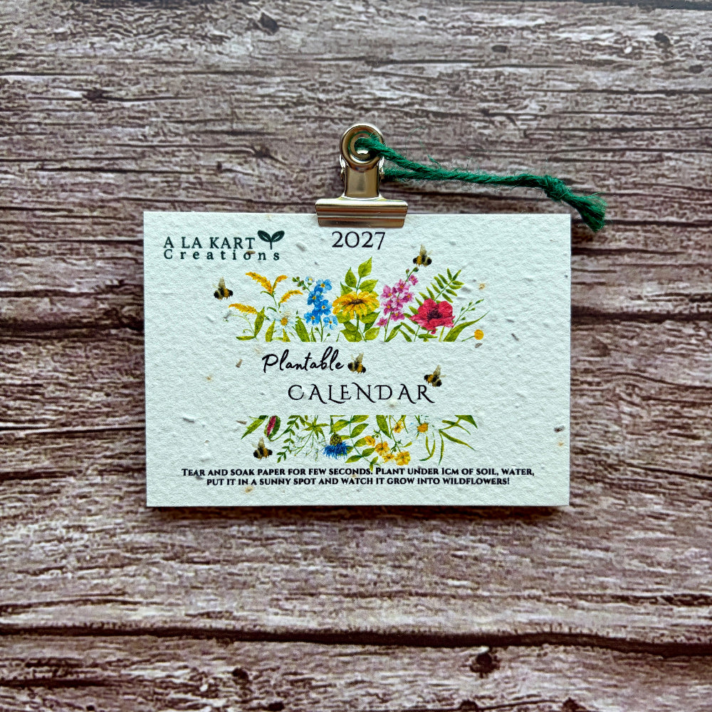 A 2027 plantable calendar card by A La Kart Creations, held by a gold clip with a green string on a rustic wooden background.