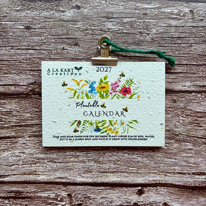 A 2027 plantable calendar card by A La Kart Creations, held by a gold clip with a green string on a rustic wooden background.