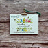 A 2027 plantable calendar card by A La Kart Creations, held by a gold clip with a green string on a rustic wooden background.
