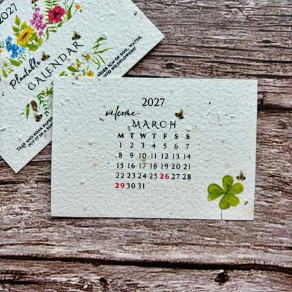 A handmade seed paper 2027 monthly calendar card with watercolor floral and butterfly illustrations, displayed on a rustic wooden background next to another calendar page.