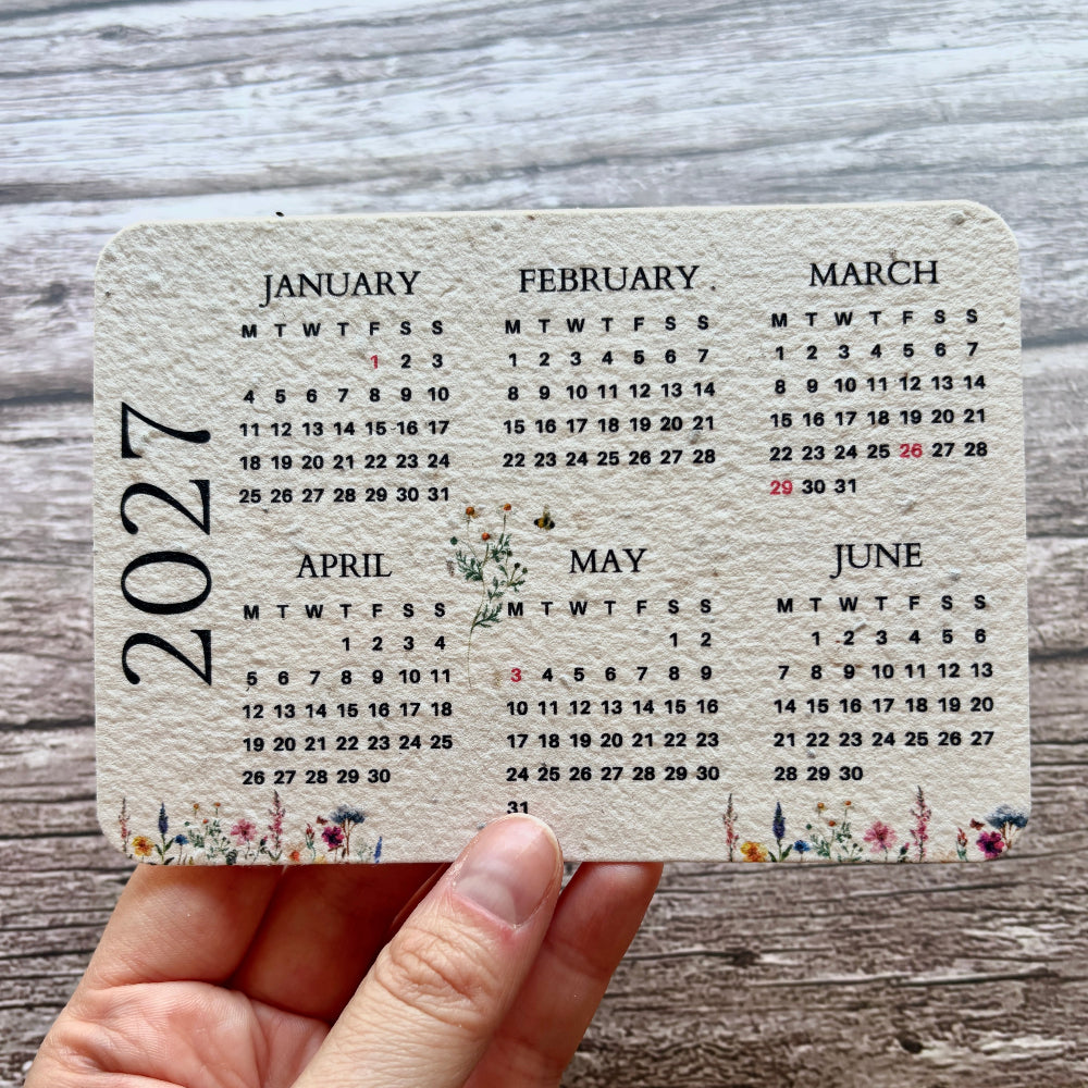 A hand holding a rectangular monthly plantable calendar card for 2027, showing the months in a grid format with small floral details along the bottom, set against a rustic gray wood background.