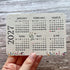A hand holding a rectangular monthly plantable calendar card for 2027, showing the months in a grid format with small floral details along the bottom, set against a rustic gray wood background.