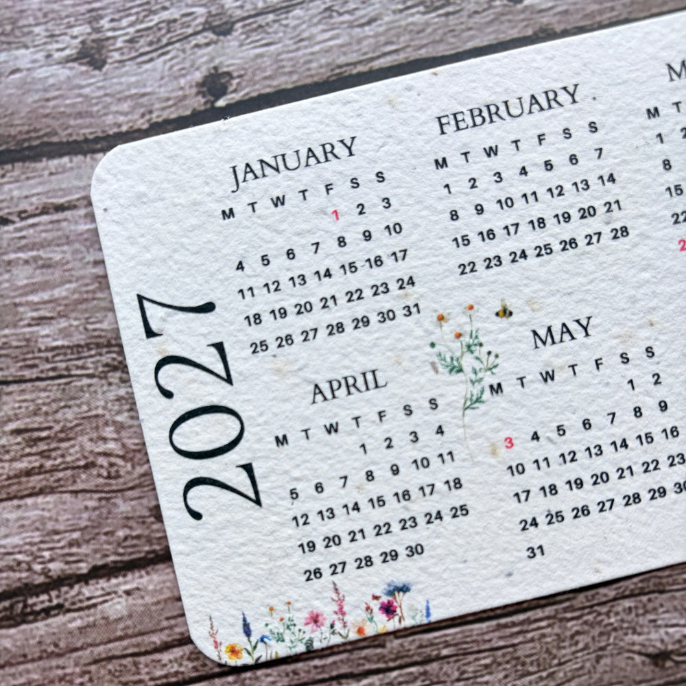 A close-up of a white seed paper calendar page showing the months of January through May 2027, printed with small floral illustrations, placed on a rustic wooden surface.