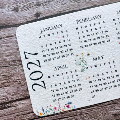 A close-up of a white seed paper calendar page showing the months of January through May 2027, printed with small floral illustrations, placed on a rustic wooden surface.