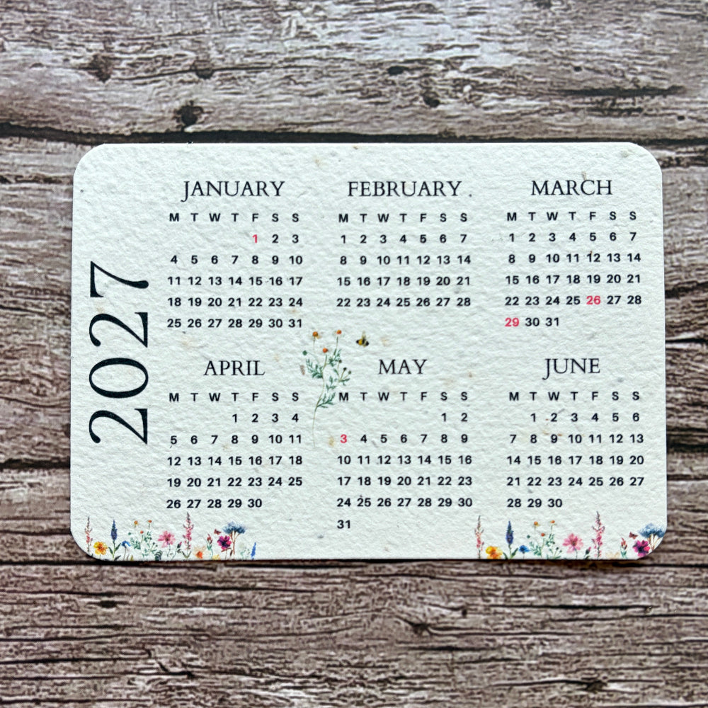 Plantable calendar card or coaster with rounded corners, printed with a 2027 monthly calendar layout and small floral illustrations along the edges, placed on a rustic weathered wood surface.
