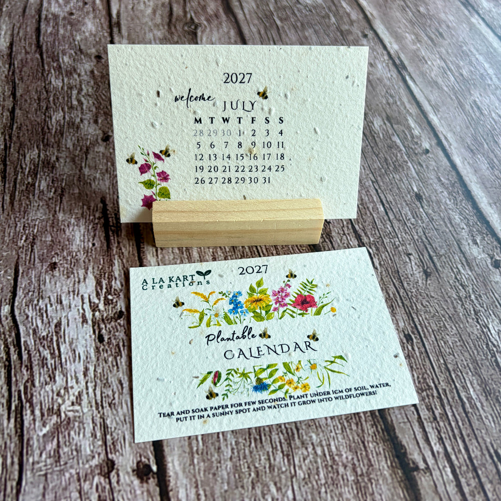 A handmade seed paper 2027 monthly calendar card set displayed on a rustic wooden surface, featuring one card standing upright on a small wooden block and another laid flat with floral illustrations.