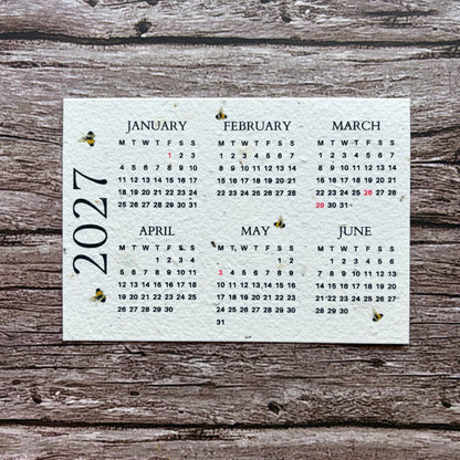 A square white seed paper calendar page showing the months of 2027 in black text, placed on a rustic weathered wood surface.