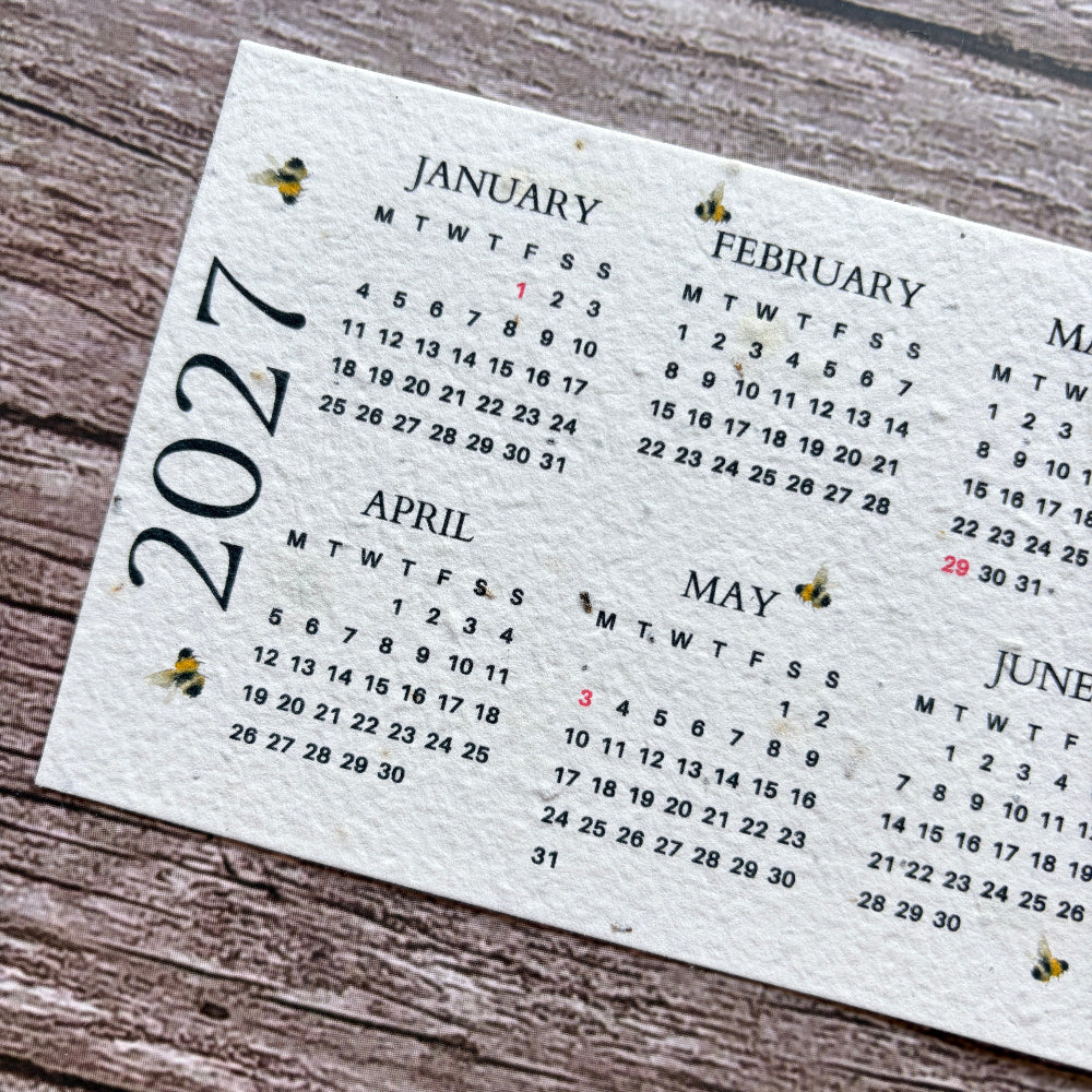 A close-up of a printed 2027 monthly calendar page with bee illustrations, placed on a rustic wooden surface.