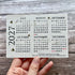 A hand holding a square, textured off-white seed paper calendar card for 2027, decorated with small bumblebees and a small plant illustration, set against a rustic gray wood plank background.