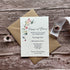 A seed paper evening wedding invitation with a floral design and text details, placed on a rustic wooden background with a small bouquet of dried flowers and a kraft envelope.