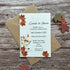 A wedding invitation card with autumn floral illustrations and leaf accents, placed on top of a brown kraft envelope over a rustic wood plank background, with decorative paper leaves around it.