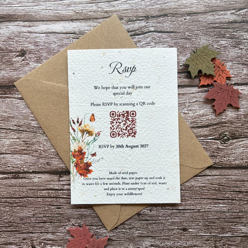 A handmade seed paper evening invitation card with a QR code and floral illustration, placed on top of a brown kraft envelope on a rustic wood surface with decorative autumn leaves.