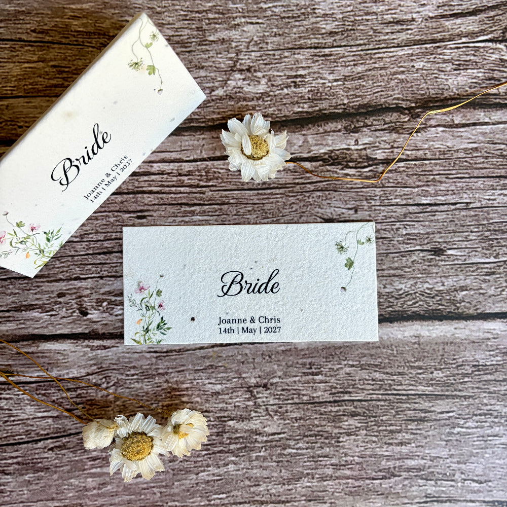 Two plantable wedding place cards with floral designs on a wooden surface with dried flowers.